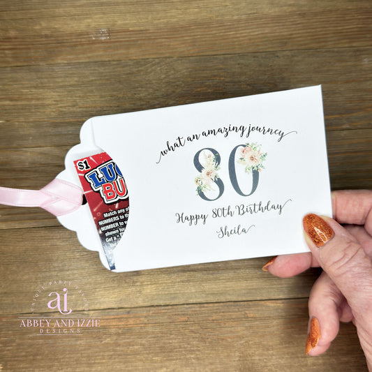 Milestone 80th Birthday Party Favors, white envelopes adorned with 80 in a floral design, personalized for the guest of honor. Slide a lottery ticket inside for a fun favor. Your choice of ribbon color, white, pink or black.