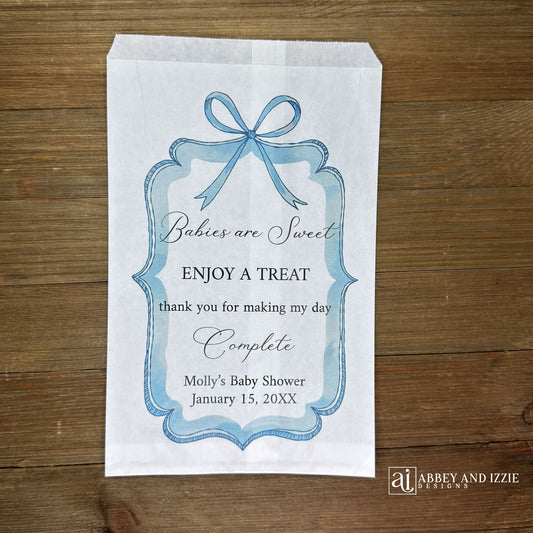 Boy baby shower favor bags adorned with a blue bow and frame printed on white paper bags. Each is personalized with 'Babies are Sweet enjoy a treat, thank you for making my day complete'. Mother to be's name and event date also printed.