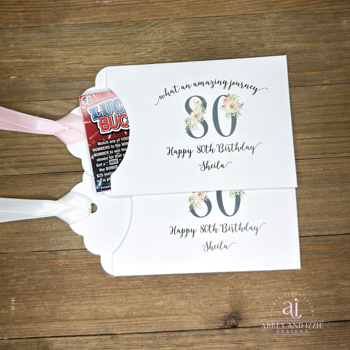 Milestone 80th Birthday Party Favors,  white envelopes adorned with 80 in a floral design, personalized for the guest of honor.  Slide a lottery ticket inside for a fun favor.  Your choice of ribbon color, white, pink or black.