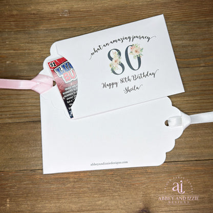Milestone 80th Birthday Party Favors,  white envelopes adorned with 80 in a floral design, personalized for the guest of honor.  Slide a lottery ticket inside for a fun favor.  Your choice of ribbon color, white, pink or black.