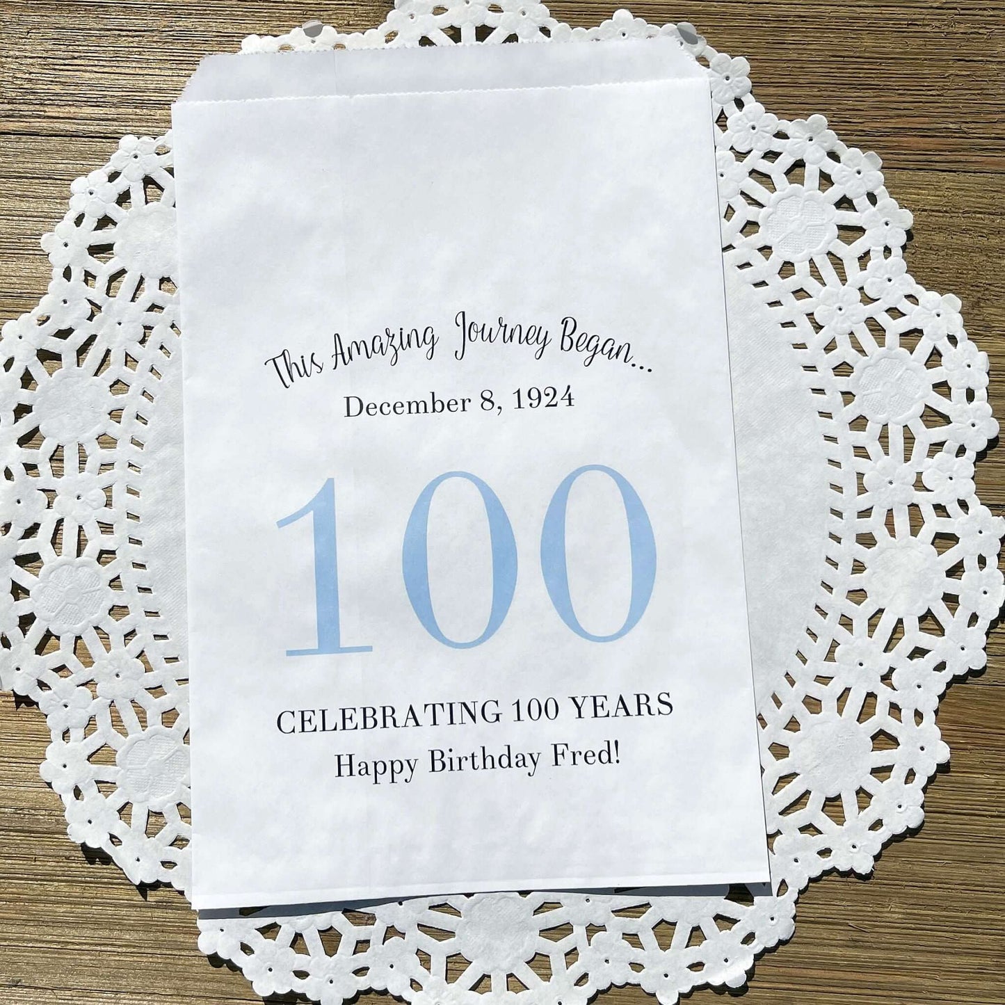 Personalized 100th Birthday Party Favor Bags, each with the name of the honoree and birth date printed.  Bags are white with a large 100 in color printed in the center.  Bags are larger than most and can hold hold cookies, candy, popcorn, goodies or even napkins and utensils.  