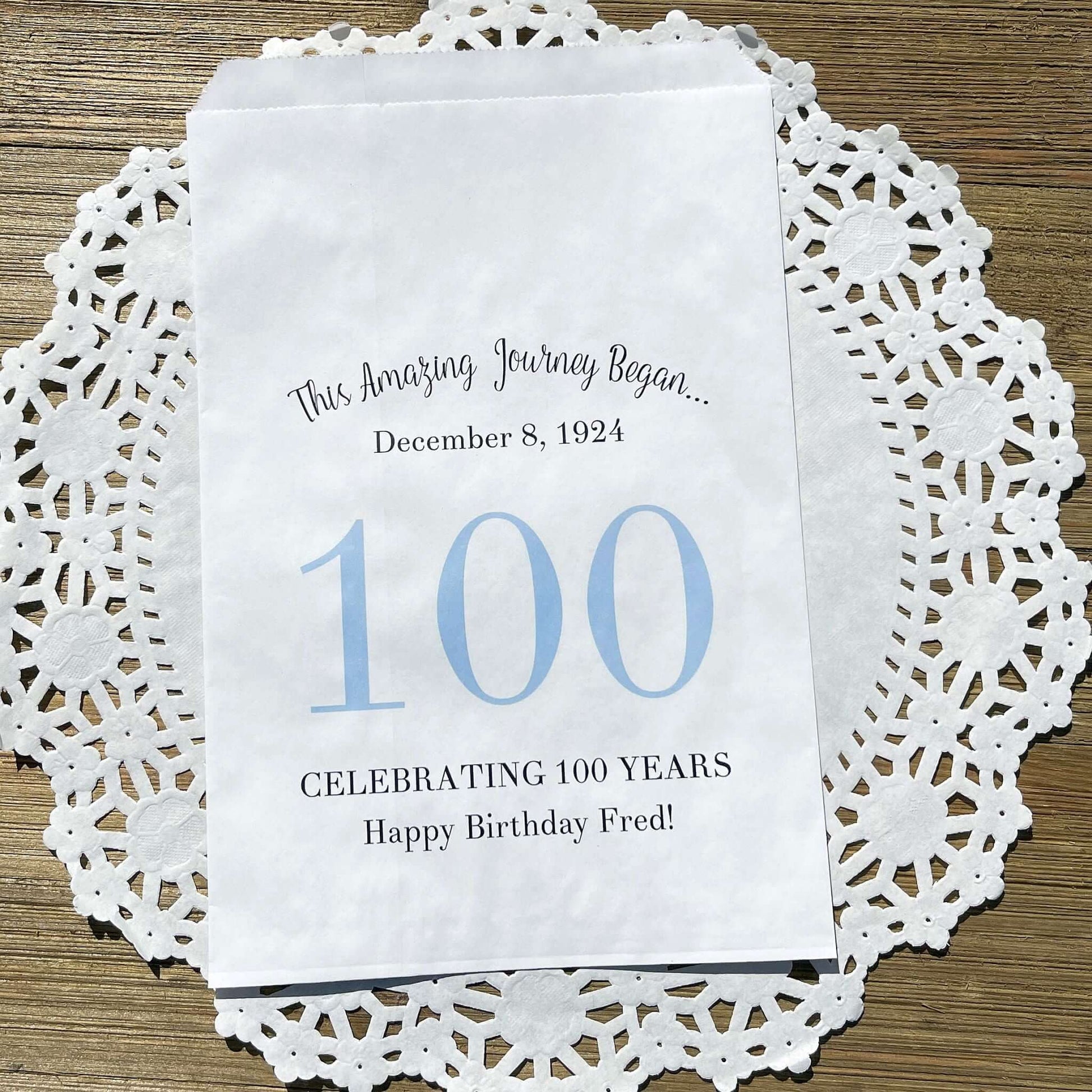 Personalized 100th Birthday Party Favor Bags, each with the name of the honoree and birth date printed.  Bags are white with a large 100 in color printed in the center.  Bags are larger than most and can hold hold cookies, candy, popcorn, goodies or even napkins and utensils.  