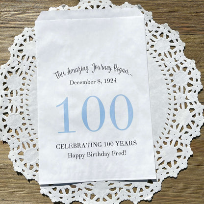 Personalized 100th Birthday Party Favor Bags, each with the name of the honoree and birth date printed.  Bags are white with a large 100 in color printed in the center.  Bags are larger than most and can hold hold cookies, candy, popcorn, goodies or even napkins and utensils.  