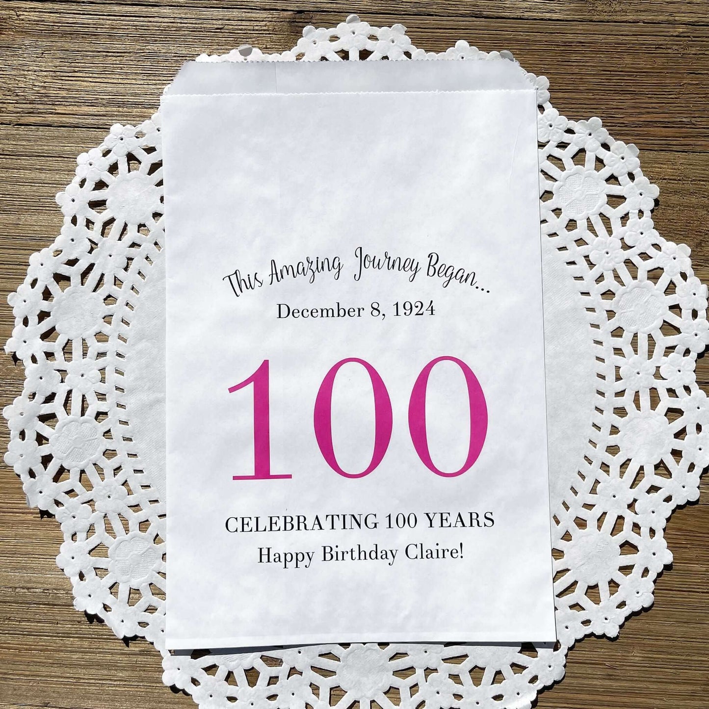 Personalized 100th Birthday Party Favor Bags, each with the name of the honoree and birth date printed.  Bags are white with a large 100 in color printed in the center.  Bags are larger than most and can hold hold cookies, candy, popcorn, goodies or even napkins and utensils.  