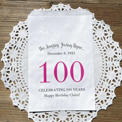 Personalized 100th Birthday Party Favor Bags, each with the name of the honoree and birth date printed.  Bags are white with a large 100 in color printed in the center.  Bags are larger than most and can hold hold cookies, candy, popcorn, goodies or even napkins and utensils.  