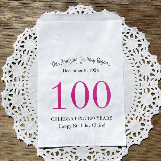 Personalized 100th Birthday Party Favor Bags, each with the name of the honoree and birth date printed.  Bags are white with a large 100 in color printed in the center.  Bags are larger than most and can hold hold cookies, candy, popcorn, goodies or even napkins and utensils.  