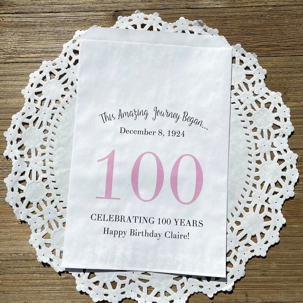 Personalized 100th birthday party favor bags, each with the name of the honoree and birth date printed.  Bags are white with a large 100 in color printed in the center.  Bags are larger than most and can hold hold cookies, candy, popcorn, goodies or even napkins and utensils. 