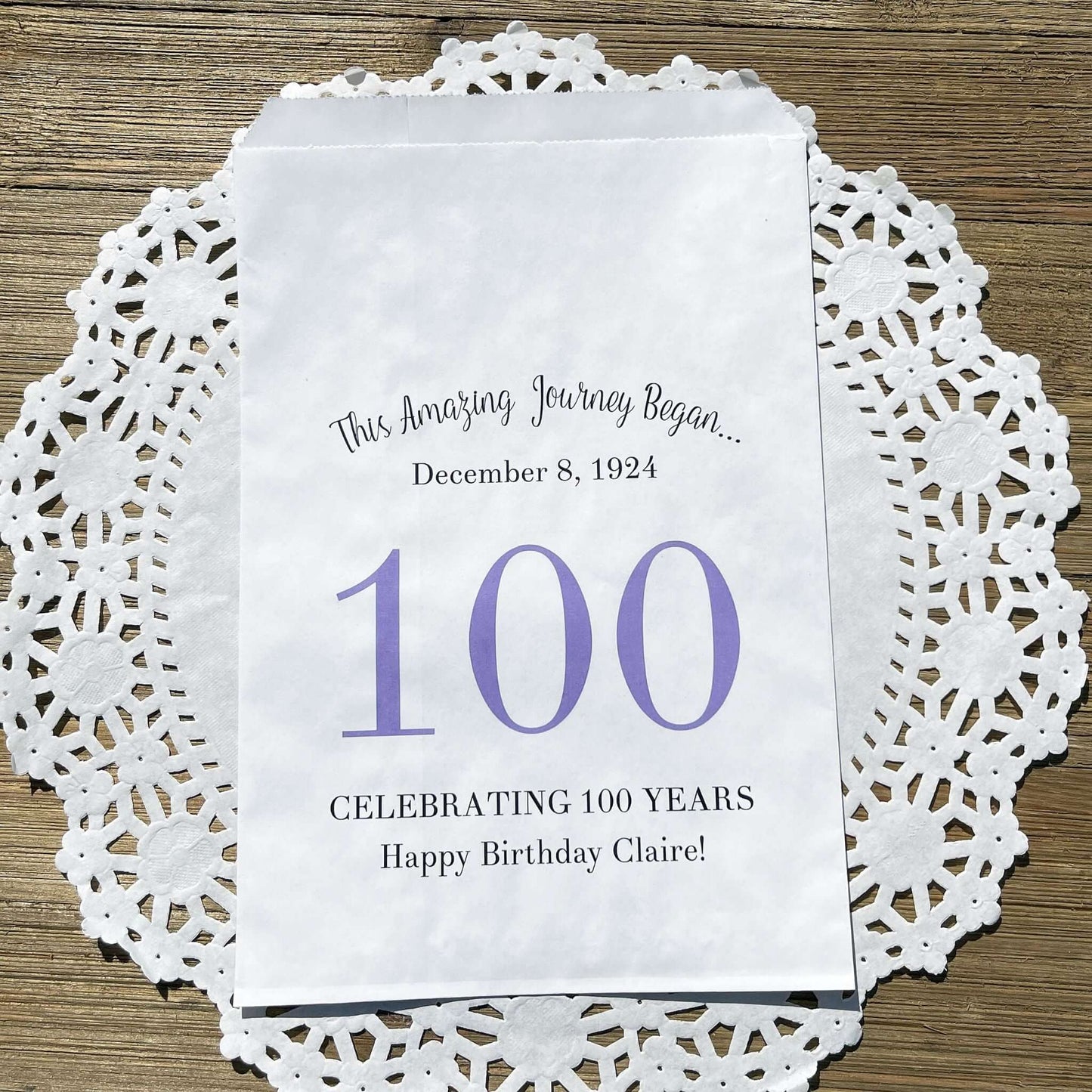 Personalized 100th Birthday Party Favor Bags, each with the name of the honoree and birth date printed.  Bags are white with a large 100 in color printed in the center.  Bags are larger than most and can hold hold cookies, candy, popcorn, goodies or even napkins and utensils.  
