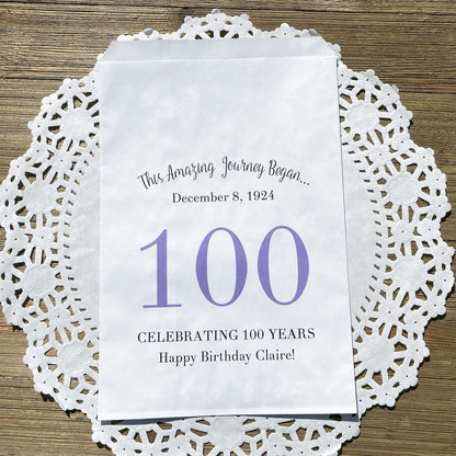 Personalized 100th Birthday Party Favor Bags, each with the name of the honoree and birth date printed.  Bags are white with a large 100 in color printed in the center.  Bags are larger than most and can hold hold cookies, candy, popcorn, goodies or even napkins and utensils.  