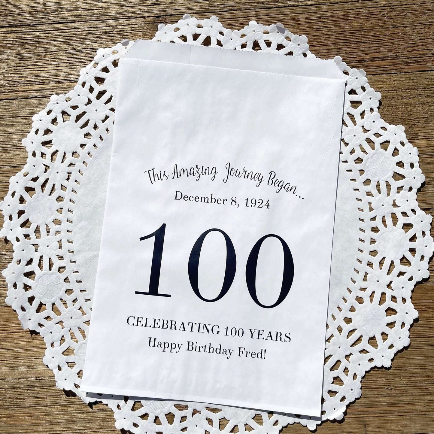 Personalized 100th Birthday Party Favor Bags, each with the name of the honoree and birth date printed.  Bags are white with a large 100 in color printed in the center.  Bags are larger than most and can hold hold cookies, candy, popcorn, goodies or even napkins and utensils.  