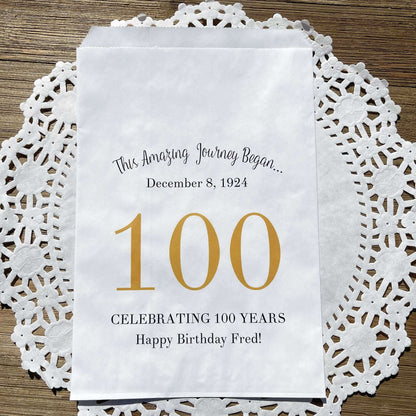 Personalized 100th Birthday Party Favor Bags, each with the name of the honoree and birth date printed.  Bags are white with a large 100 in color printed in the center.  Bags are larger than most and can hold hold cookies, candy, popcorn, goodies or even napkins and utensils.  