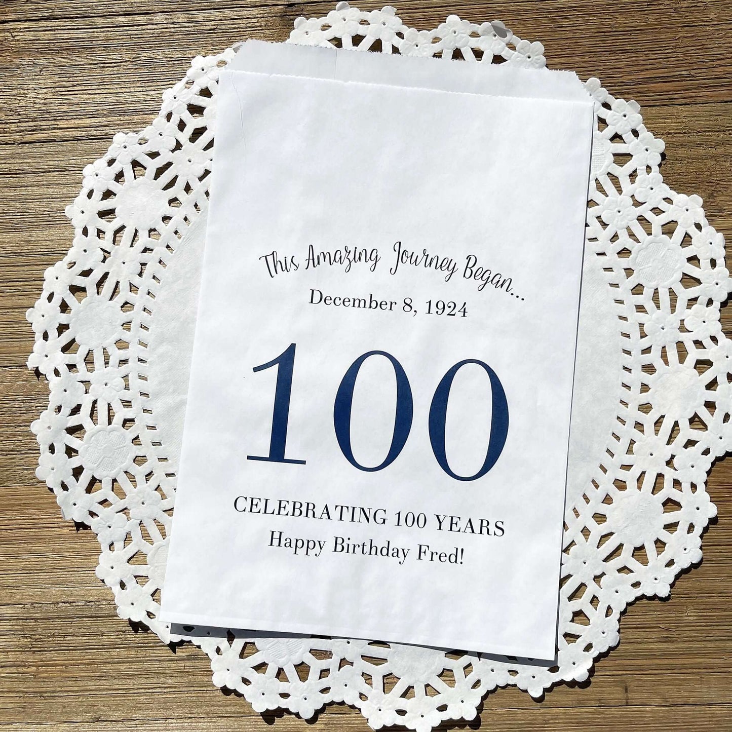 Personalized 100th Birthday Party Favor Bags, each with the name of the honoree and birth date printed.  Bags are white with a large 100 in color printed in the center.  Bags are larger than most and can hold hold cookies, candy, popcorn, goodies or even napkins and utensils.  