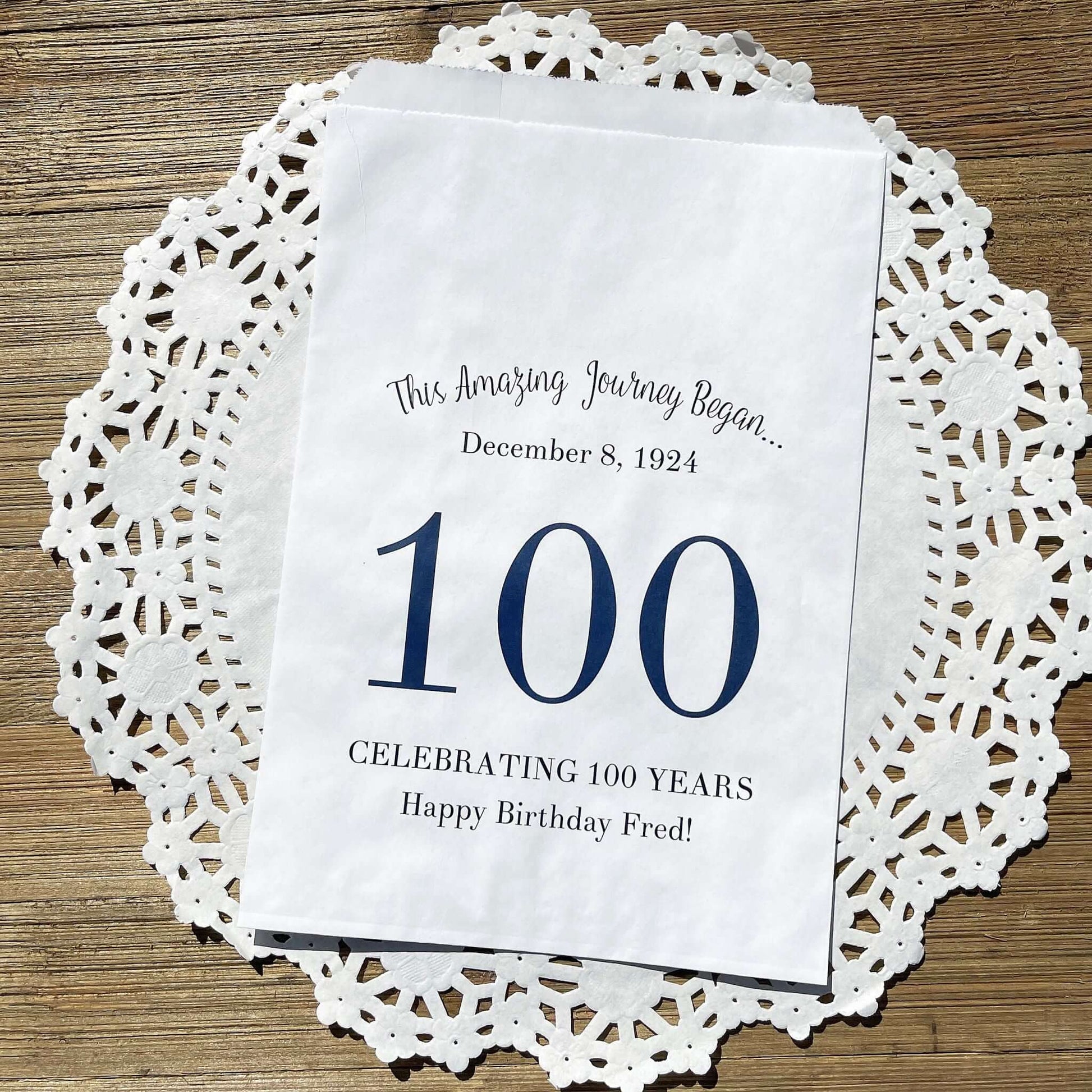 Personalized 100th Birthday Party Favor Bags, each with the name of the honoree and birth date printed.  Bags are white with a large 100 in color printed in the center.  Bags are larger than most and can hold hold cookies, candy, popcorn, goodies or even napkins and utensils.  