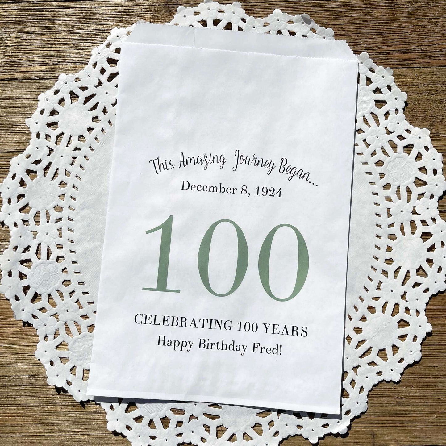 Personalized 100th Birthday Party Favor Bags, each with the name of the honoree and birth date printed.  Bags are white with a large 100 in color printed in the center.  Bags are larger than most and can hold hold cookies, candy, popcorn, goodies or even napkins and utensils.  
