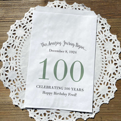 Personalized 100th Birthday Party Favor Bags, each with the name of the honoree and birth date printed.  Bags are white with a large 100 in color printed in the center.  Bags are larger than most and can hold hold cookies, candy, popcorn, goodies or even napkins and utensils.  