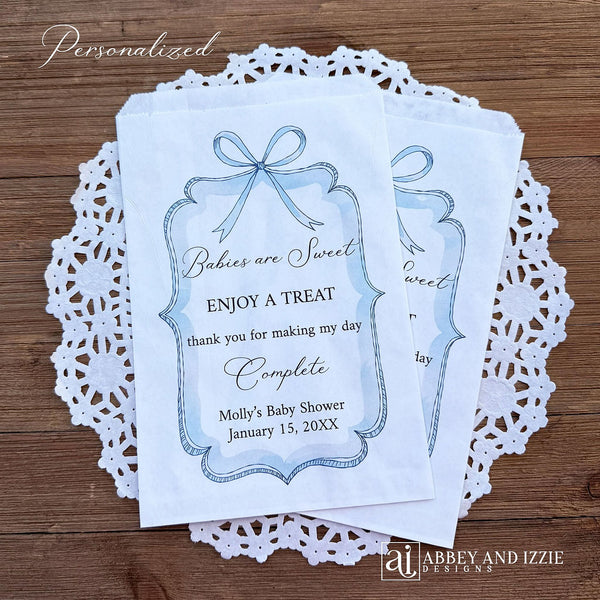Boy baby shower favor bags adorned with a blue bow and frame printed on white paper bags.  Each is personalized with 'Babies are Sweet enjoy a treat, thank you for making my day complete'.  Mother to be's name and event date also printed.  