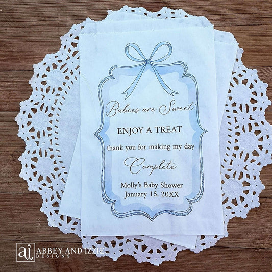 Boy baby shower favor bags adorned with a blue bow and frame printed on white paper bags.  Each is personalized with 'Babies are Sweet enjoy a treat, thank you for making my day complete'.  Mother to be's name and event date also printed.  