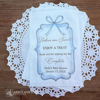 Boy baby shower favor bags adorned with a blue bow and frame printed on white paper bags.  Each is personalized with 'Babies are Sweet enjoy a treat, thank you for making my day complete'.  Mother to be's name and event date also printed.  