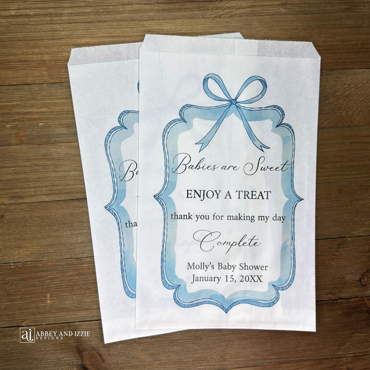Boy baby shower favor bags adorned with a blue bow and frame printed on white paper bags.  Each is personalized with 'Babies are Sweet enjoy a treat, thank you for making my day complete'.  Mother to be's name and event date also printed.  
