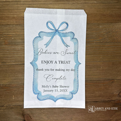Boy baby shower favor bags adorned with a blue bow and frame printed on white paper bags.  Each is personalized with 'Babies are Sweet enjoy a treat, thank you for making my day complete'.  Mother to be's name and event date also printed.  