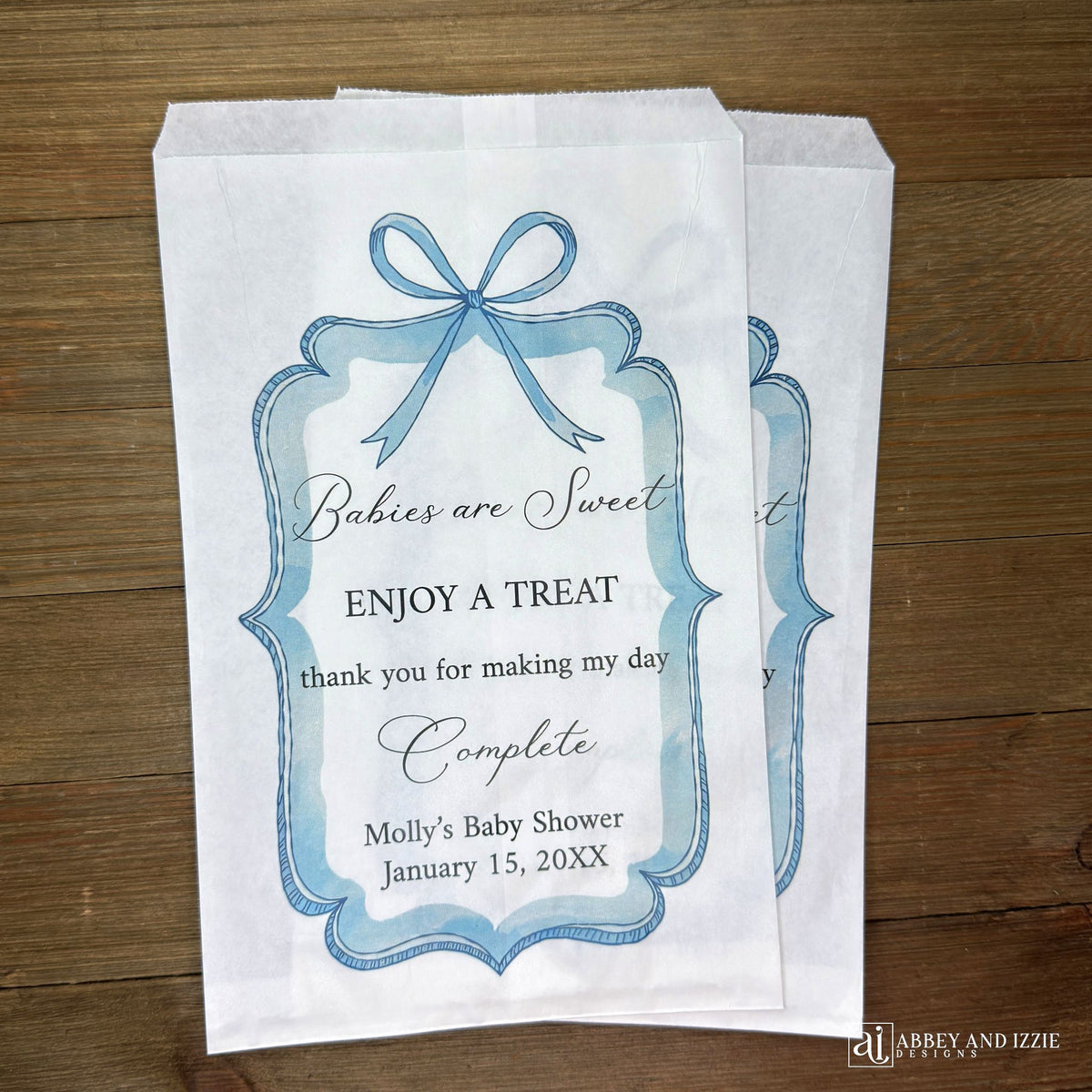 Boy baby shower favor bags adorned with a blue bow and frame printed on white paper bags.  Each is personalized with 'Babies are Sweet enjoy a treat, thank you for making my day complete'.  Mother to be's name and event date also printed.  