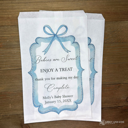 Boy baby shower favor bags adorned with a blue bow and frame printed on white paper bags.  Each is personalized with 'Babies are Sweet enjoy a treat, thank you for making my day complete'.  Mother to be's name and event date also printed.  