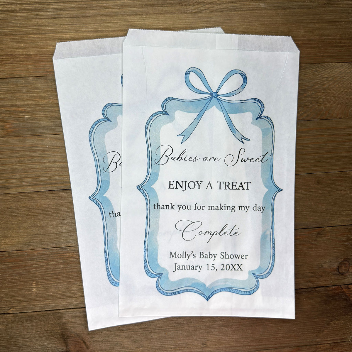 Boy baby shower favor bags adorned with a blue bow and frame printed on white paper bags.  Each is personalized with 'Babies are Sweet enjoy a treat, thank you for making my day complete'.  Mother to be's name and event date also printed.  