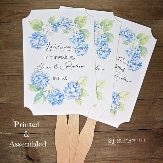 Elevate your wedding ambiance with our personalized blue hydrangea fans, beautifully crafted to complement your special day.  Printed on white card stock with 'welcome to our wedding' and the bride and grooms names with wedding date printed. 