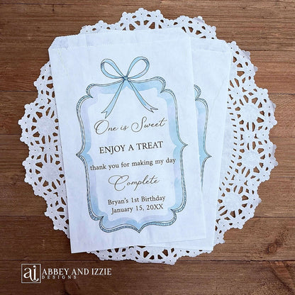 Personalized first birthday party favor bags, 'one is sweet, enjoy a treat, thank you for making my day complete'.  Printed on white paper bags and adorneed with a blue bow and border these will add a touch of elegance to the treat table. 