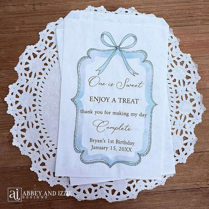 Personalized first birthday party favor bags, 'one is sweet, enjoy a treat, thank you for making my day complete'.  Printed on white paper bags and adorneed with a blue bow and border these will add a touch of elegance to the treat table. 