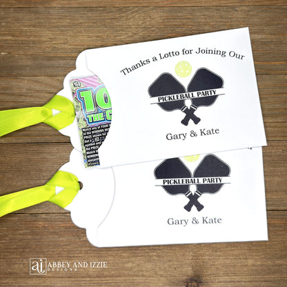 Personalized Pickleball Party Favors that will be fun for all your guests. Adonred with a pickleball and paddles these are personalized for the event. Printed on white cardstock with green ribbon attached. Slide a lottery ticket in the open end and see who wins.