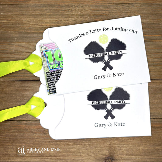 Personalized Pickleball Party Favors that will be fun for all your guests. Adonred with a pickleball and paddles these are personalized for the event. Printed on white cardstock with green ribbon attached. Slide a lottery ticket in the open end and see who wins.