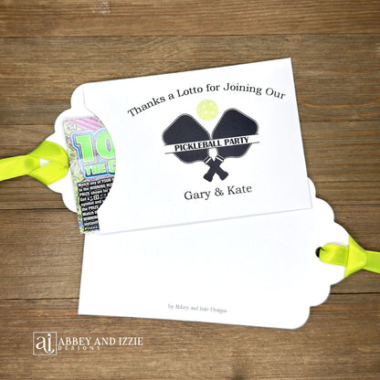 Personalized Pickleball Party Favors that will be fun for all your guests. Adonred with a pickleball and paddles these are personalized for the event. Printed on white cardstock with green ribbon attached. Slide a lottery ticket in the open end and see who wins.