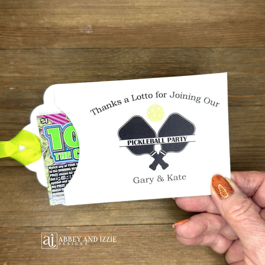 Personalized Pickleball Party Favors that will be fun for all your guests. Adonred with a pickleball and paddles these are personalized for the event. Printed on white cardstock with green ribbon attached. Slide a lottery ticket in the open end and see who wins.