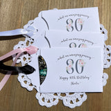 Lottery ticket envelopes for 80th birthday party favors. White envelopes adorned with 80 in a floral design, personalized for the guest of honor.