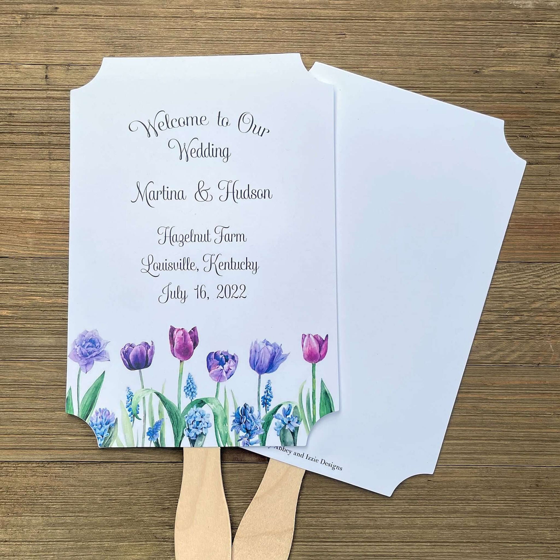 Tulip Wedding Guest Fans adorned with purple tulips.  Fans are printed on white card stock, two sided with handle hidden between, printing on one side only.  Printing included bride and groom, place, city and state and date.  Shipped assembled with wooden handle attached.