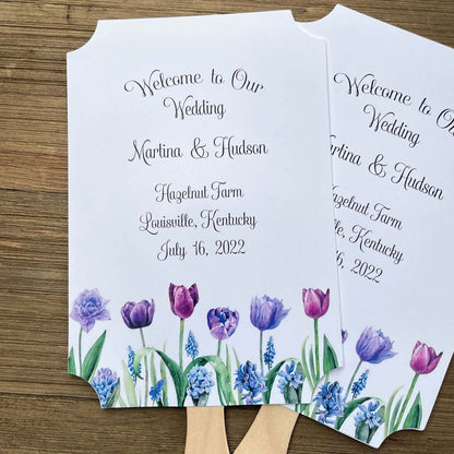 Tulip Wedding Guest Fans adorned with purple tulips.  Fans are printed on white card stock, two sided with handle hidden between, printing on one side only.  Printing included bride and groom, place, city and state and date.  Shipped assembled with wooden handle attached.