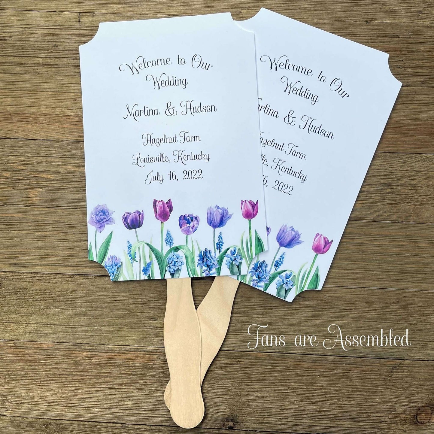 Tulip Wedding Guest Fans adorned with purple tulips.  Fans are printed on white card stock, two sided with handle hidden between, printing on one side only.  Printing included bride and groom, place, city and state and date.  Shipped assembled with wooden handle attached.